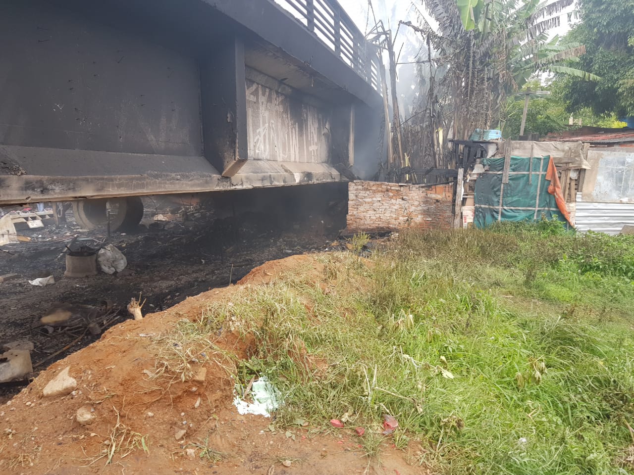 Terreno incendiado embaixo de viaduto, paredes queimadas, barraco ao fundo e uma parte de terreno com gramado 